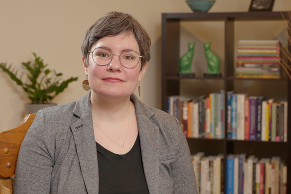 Woman with short brown hair, glasses, and a grey blazer looks at the camera with bookshelf and plants behind