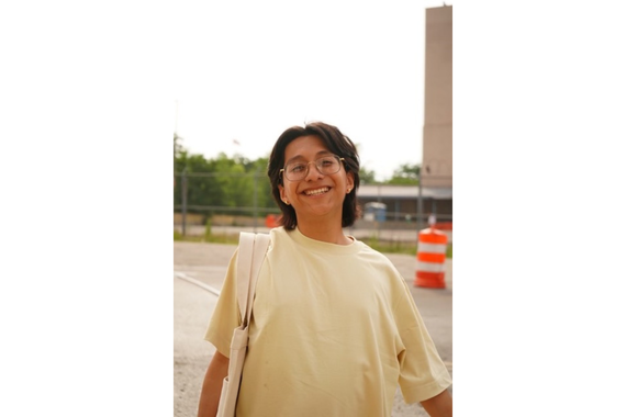 Portrait of a person with dark hair to shoulders, wearingthinly framed gold glasses and a light yellow t-shirt, smiling.