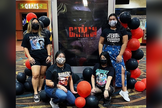 Four women wearing Robert Pattinson shirts and standing in front of The Batman movie poster at a movie theater