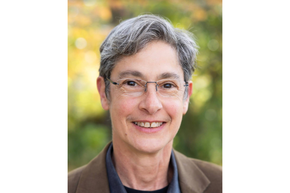 Portrait of a person with short grey hair, wearing thinly framed glasses, smiling. The person is standing in front of a green background while wearing a brown blazer and blue collared shirt. 