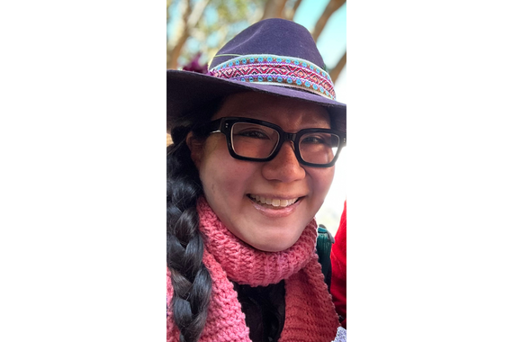 Portrait of a woman wearing black framed glasses and a grey hat with a short brim, smiling. The person is wearing a pink scarf wrapped around their neck and their hair is dark brown in a braid on their right side. 