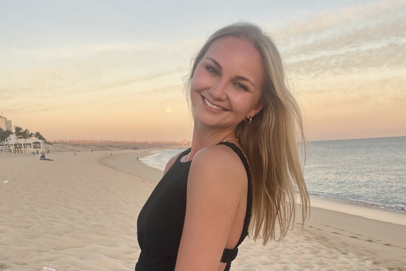 A woman smiles at the camera with a beach and sunset background