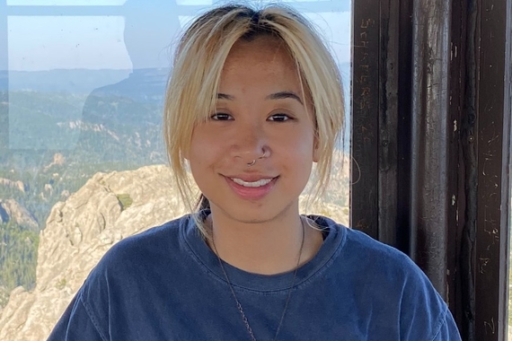 An Asian person with tan skin; short blond hair with black roots; thick blue shirt; and silver necklace, smiling and standing in front of a window that showcases a mountainous, grassy landscape