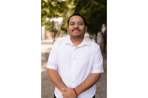 Person with short brown hair and a brown mustache standing with hands joined together smiling slightly. They are wearing a short sleeve white collared polo shirt and they are standing in front of a paved path with trees in the background.