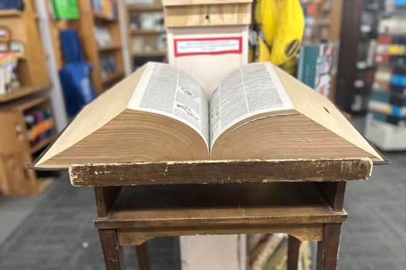 A corduroy bound dictionary weighing over 25 pounds and measuring 9 inches tall sits on a rickety wooden end table in a bookstore.