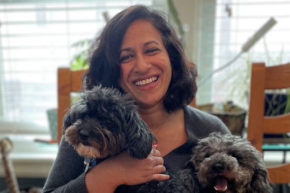 Head and shoulders of person with brown hair to shoulders and  brown skin, wearing grey top, smiling, and holding two grey fluffy dogs; windows behind