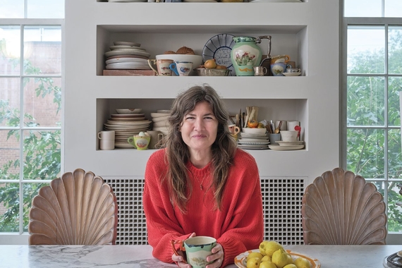 Ginny Sims in red sits with a coffee mug in front of shelves full of ceramics