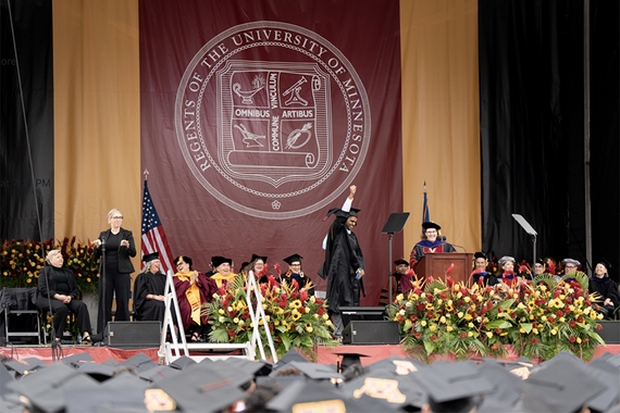 People in black robes on stage with gold flowers to side, maroon banner in background with white seal; grad caps in foreground