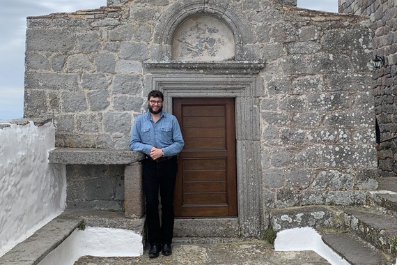 Alex Magnolia, a young man with a black beard and black hair, stands in a thousand-year-old stone monastery 