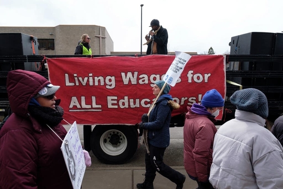 A group of people wearing coats and hats carry signs supporting striking teachers. Behind them is a truck with a banner that reads Living Wages for All Educators. A person stands in the back of the truck with a microphone.