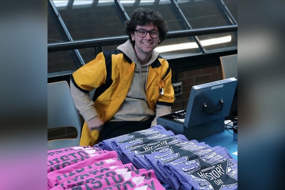 Tyler Bradley, a young man with black hair and a yellow coat, sits at a table selling History Day shirts