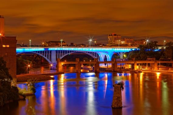 The new I-35 bridge illuminated light blue above the Mississippi at night, Moos Tower and other University buildings behind it 