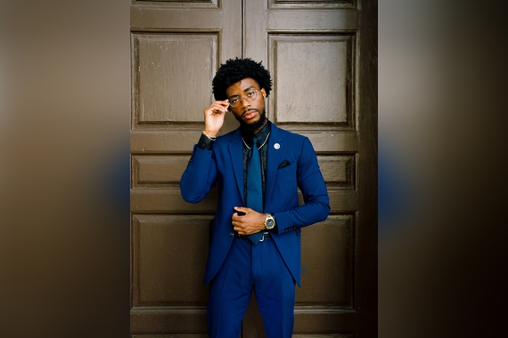 Alum Ighedosa Ogbeide wears a blue suit while standing in front of a wooden backdrop.