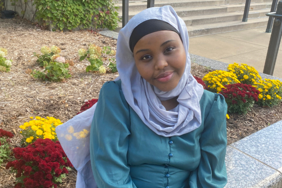 Ilhan Adan wearing a blue hijab and shirt, sitting outside in front of a flower garden on a clear day. 
