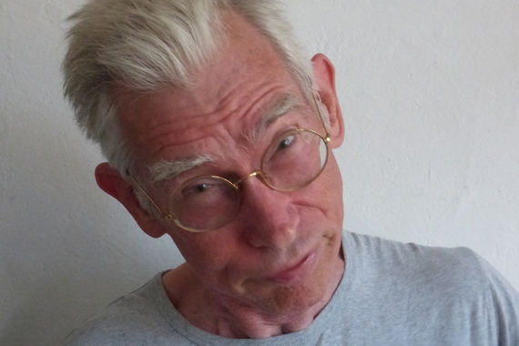 Head and shoulders of person with short white hair and light skin, tilting head left and wearing wire-rimmed glasses and grey t-shirt
