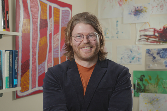 Joseph Mullins, a man with a beard, moustache, and glasses wearing a black blazer, smiles at the camera in a vibrant office