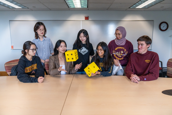Students in maroon and gold apparel take turns rolling two oversized yellow dice.