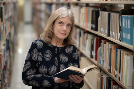 Ann Waltner stands in the library, holding an open book and smiling