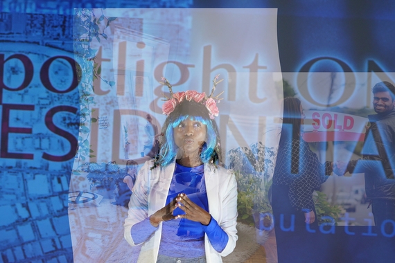 A still video image of a young black woman dressed as a millenial executive in a blue spandex turtleneck, white blazer, bright blue and black wig, and a flower crown. She stands in front of a projected stage screen that says "Spotlight on Residential" and a digital image of a wine picnic floats in front of her.