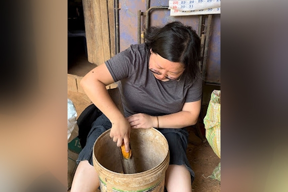 Woman with black hair, gray shirt, and glasses sitting down and using tool to shred corn in tan bucket.