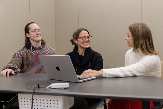 Sat at a long table, students Alana Miller (right) and Ethan Schmid (left)and Professor Lisa Hilbink between them are talking and smiling. They're surrounding a laptop atop the table, the Alana Miller with their hand on the trackpad.