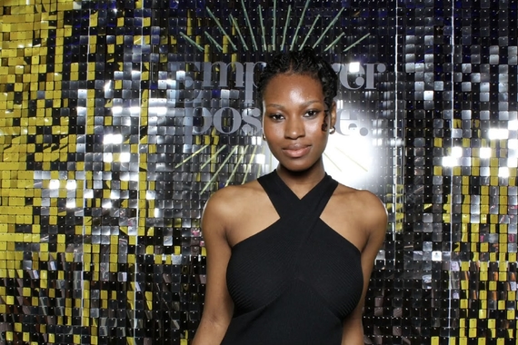 A black woman with black braids in a bun wearing a black dress, smiling in front of a black background with gold sequences