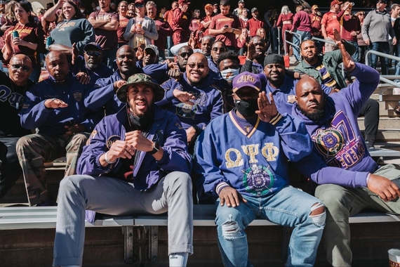 A group of Black men of different ages wearing purple Omega Psi Phi hats, shirts, and jackets sitting on bleachers at an athletic event