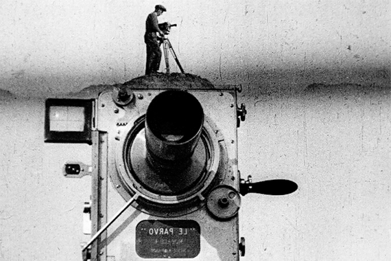 Grainy film still of a small man standing on a giant camera