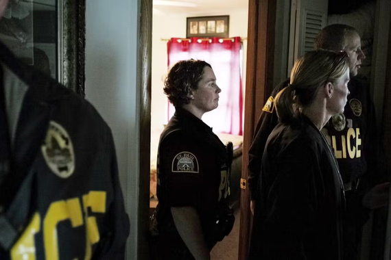 A police officer stands in a doorway looking toward the right. Other officers look in the same direction