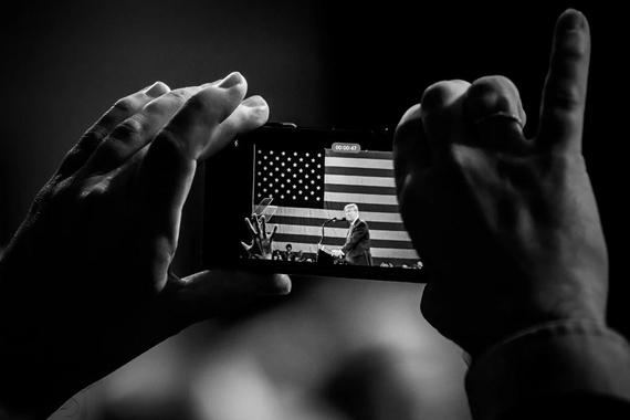 Two hands hold up a cellphone capturing an image of Donald Trump standing in front of the American flag.
