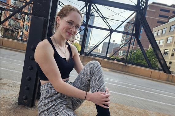 Person sitting against metal post with one knee up, light brown hair drawn back, light skin, wearing glasses and sleeveless black top and black and white plaid pants; black metal structure in background and city buildings