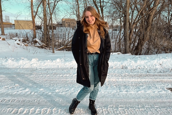 Person standing on snowy road, with light brown hair past shoulders, light skin, smiling, hands in coat pockets, wearing black coat to knees, tan turtleneck, light blue jeans, black boots, bare trees in background
