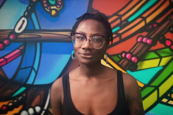 Black woman wearing glasses and a tank top smiles in front of a mural