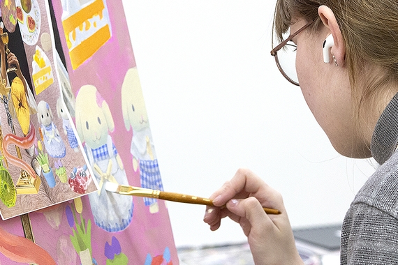 A student listens to earbuds and works on a painting of pink and white cartoon characters