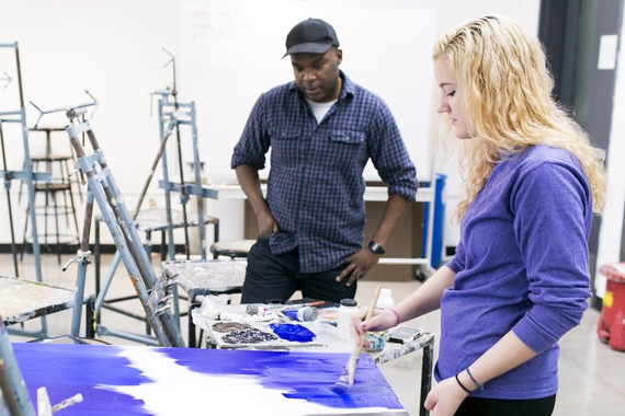 A teacher watches a student paint in shades of purple surrounded by metal easels