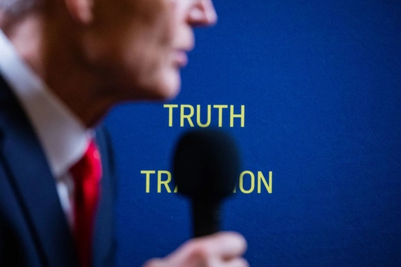Senator Rick Scott, his face out of focus and partly out of frame, holds a microphone, with the words "truth" and tradition" in yellow visible on the blue wall behind him. "Tradition" is largely blocked by the microphone.