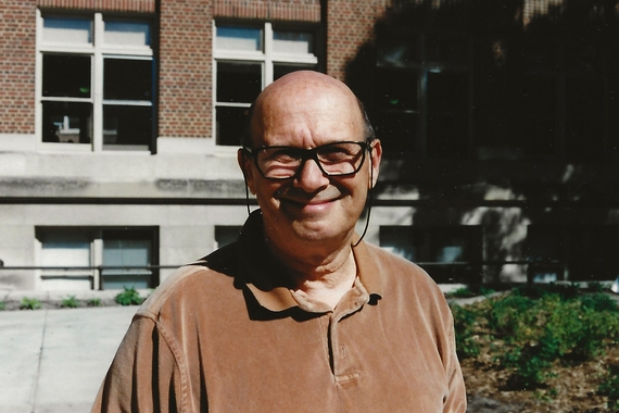 Photo of person with bald head, glasses, pale skin, wearing brown shirt, standing in front of brick building