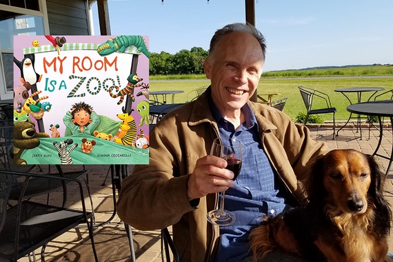 Small image of children's book MY ROOM IS A ZOO (illustration of child in bed with various animals around edge) over photograph of older person, with white short hair and light skin in brown jacket and blue shirt holding wine glass sitting behind brown dog; background is black iron chairs and tables and green field beyond