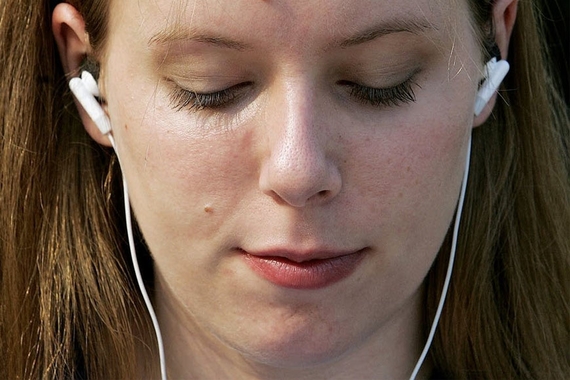 A young white woman with brown hair listens to music on wired earbuds with her eyes closed