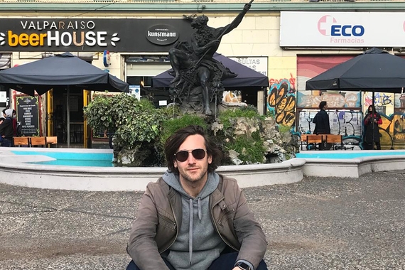 Person with dark hair and light skin, wearing sunglasses and grey jackets, sits in front of public statuary and wading pool, storefronts