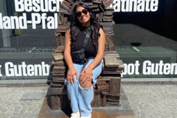 A woman with tan skin, sleeveless black sweater, ripped blue jeans, black purse, and white sneakers smiling and sitting on a metal chair outside made of books. 