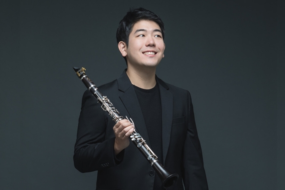 Sangyoon Kim, in a black suit, holds his clarinet against a dark background