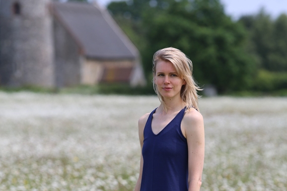 Person with blond hair to shoulders and light skin, wearing sleeveless blue top and standing in grassy field with barn and green trees in background