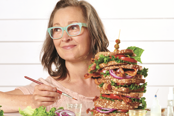 Woman with blonde hair and glasses posing behind a large sandwich