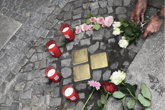 an image of several Stoplersteine, small brass plates put into the ground outside of the homes of Holocaust victims, surrouned by 4 red candles and flowers. 