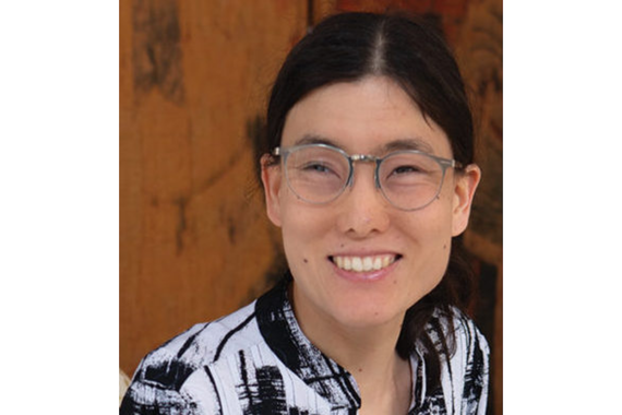 Person with dark hair tied behind their head wearing circularly framed silver glasses smiling. The person is wearing a black and white striped design shirt. 