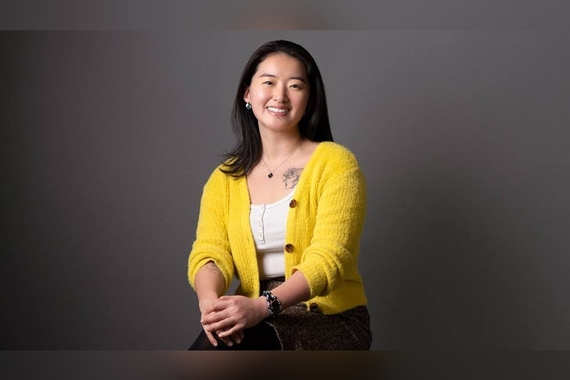 Shujianing Li wearing a yellow sweater, smiling against a black backdrop