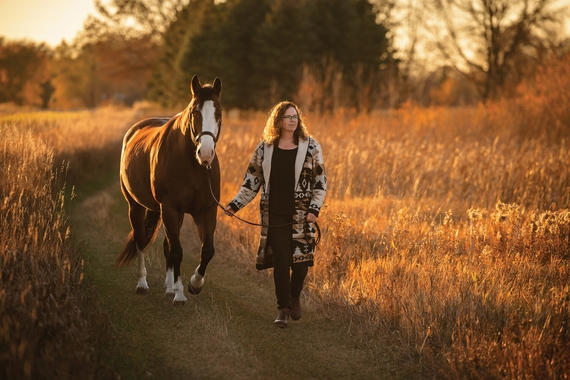Shelley Paulson walks down a path bordered by tall grasses with a horse following her.