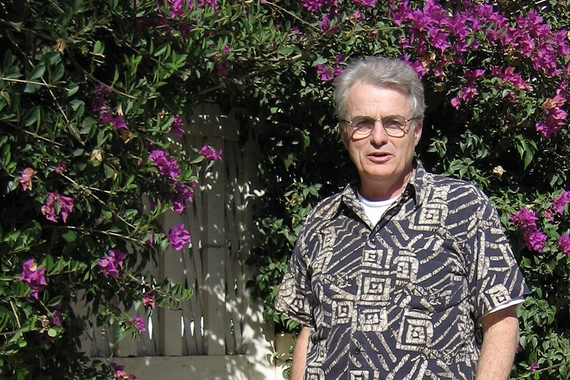 Person with short grey hair, light skin, and glasses, wearing patterned black and white shirt, standing in front of lush bush with purple flowers