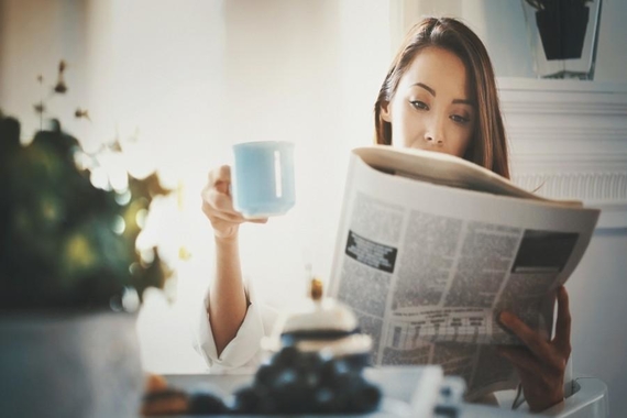 Woman sitting at a table holding a coffee mug and reading a newspaper
